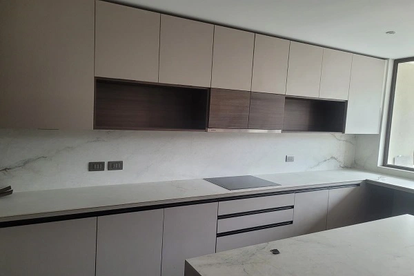 Cocina moderna fabricada en melamina en style muebles Chile con gabinetes beige y marrón, cubierta de cuarzo marmolado y repisa abierta central bajo luz natural