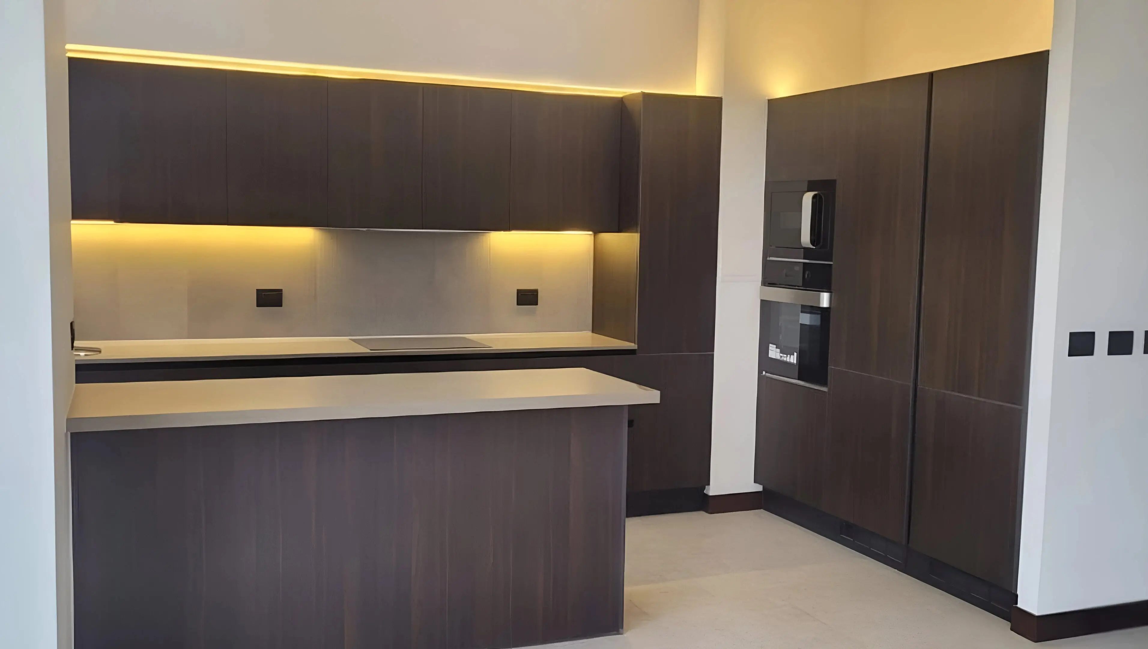 Cocina moderna con muebles en melamina oscura, encimera con cocina de inducción, iluminación cálida bajo muebles altos, horno y microondas empotrados, diseño minimalista y elegante